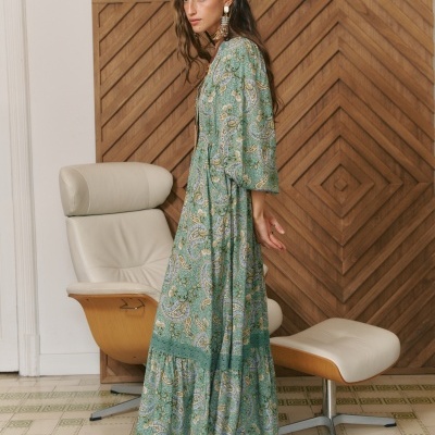 Vestido longo verde com padrão floral e estampado cachemira usado por mulher em ambiente interior com cadeira de couro bege e parede de madeira