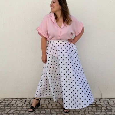 Mulher com blusa rosa clara, saia branca com bolinhas pretas e sandálias pretas em calçada de pedra