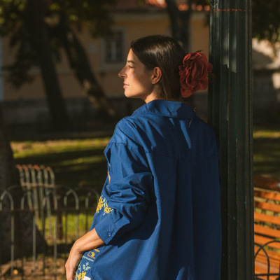Mulher com camisa azul bordada e flor vermelha no cabelo encostada a um poste verde ao ar livre