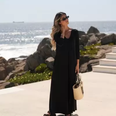 Mulher com vestido longo preto, óculos de sol e saco de palha ao ar livre junto ao mar