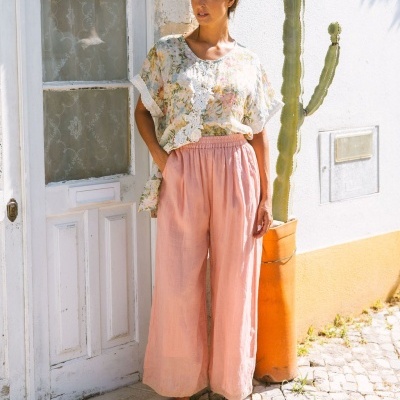Mulher com roupa leve floral e calças rosa ao lado de porta branca e cacto