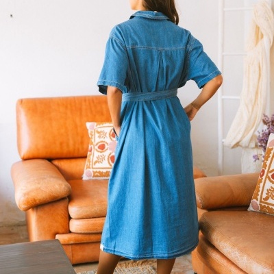 Mulher de costas a usar vestido midi azul estilo ganga com cinto, em sala com sofá castanho e almofadas decorativas.