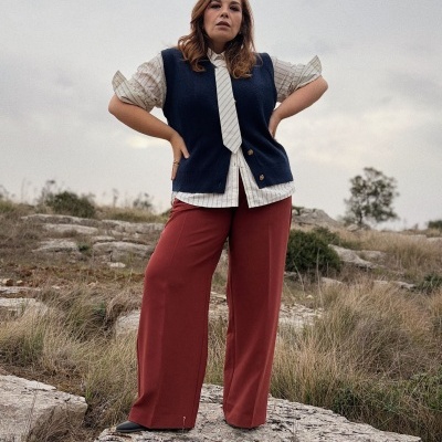 Mulher com camisa às riscas, colete azul, gravata branca, calças largas castanhas e sapatos pretos em ambiente natural rochoso.