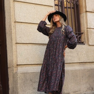 Mulher com vestido comprido floral, botas castanhas e chapéu preto em ambiente de cidade junto a prédio.