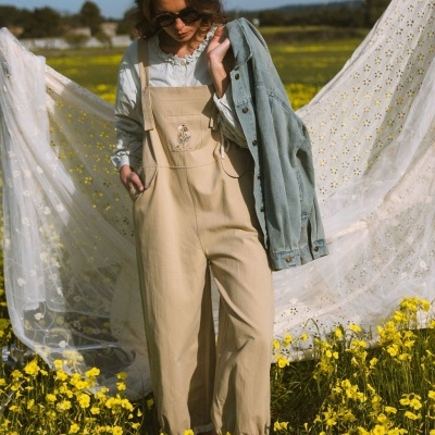Mulher com macacão bege bordado e blusa branca, casaco de ganga azul claro na mão, num campo florido amarelo com tecido rendado branco ao fundo.