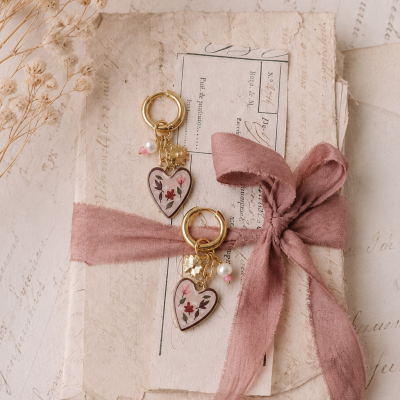 Brincos de coração com flores em fundo branco, sobre cartas antigas amarradas com fita rosa