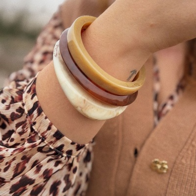 Pulseiras largas em tons creme, castanho e bege no pulso de uma mulher com blusa com padrão animal e casaco bege.