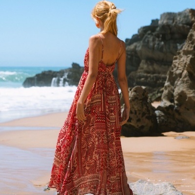 Mulher de costas a usar vestido longo vermelho e bege numa praia com pedras e mar