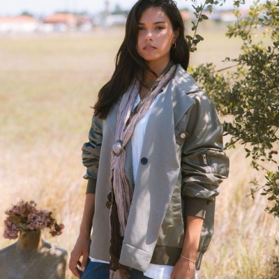 Mulher jovem com casaco bege e verde, cachecol bege claro e jeans, ao ar livre em campo seco.