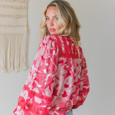 Blusa feminina vermelha e branca com padrão floral e detalhes em renda, vista de costas, com jeans azul claro e parede de fundo cinza e tapeçaria de algodão