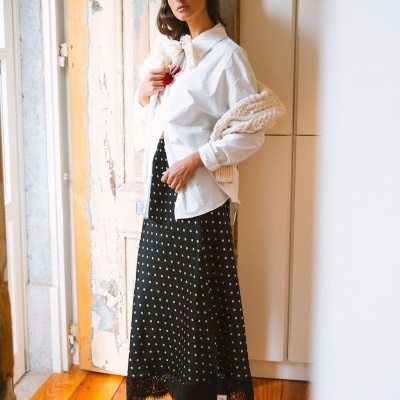 Mulher com camisa branca, saia preta com bolinhas brancas, botas pretas e casaco de malha creme em interior