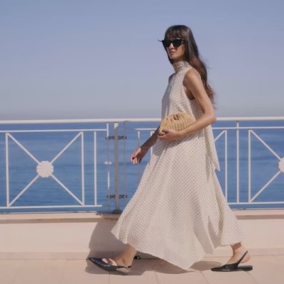 Mulher com vestido longo branco com pequenos pontos pretos e sandálias pretas, segurando mala bege ao ar livre junto ao mar