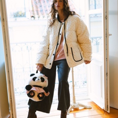 Mulher com casaco creme de pontos pretos e mala panda de peluche num interior com varanda