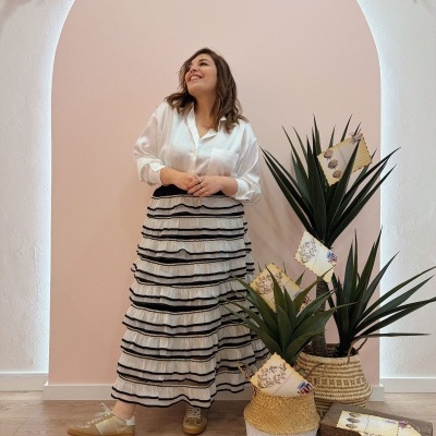 Mulher com camisa branca e saia longa branca com babados pretos em ambiente interior com plantas em vasos de cestaria.