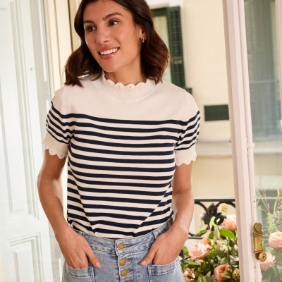 Mulher com camisola de riscas azul e branca e saia de ganga com botões à frente, junto a uma varanda com flores.