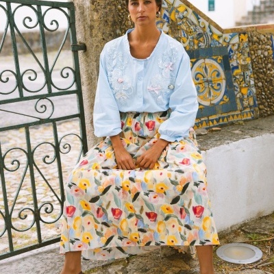 Mulher sentada com blusa azul clara bordada, saia florida e sapatos castanhos num exterior com azulejos