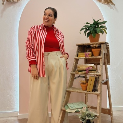 Mulher com camisa de botões vermelha e branca às riscas com calças largas bege ao lado de escada de madeira com plantas e livros.
