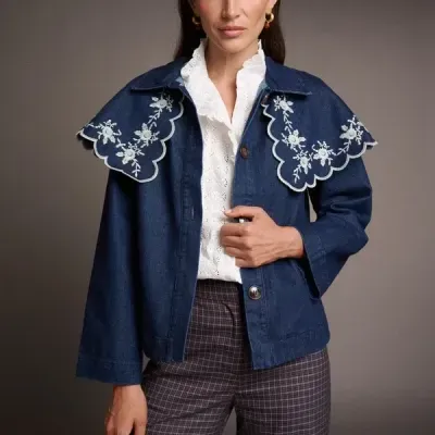 Mulher veste blusão azul tipo ganga com gola bordada, camisa branca de renda e calças aos quadrados.