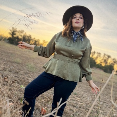Mulher com blusa verde e chapéu castanho numa paisagem de campo ao pôr do sol