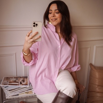 Mulher com camisa rosa listrada, calças brancas e botas castanhas, segurando smartphone e mala preta, sentada num suporte metálico com revistas.