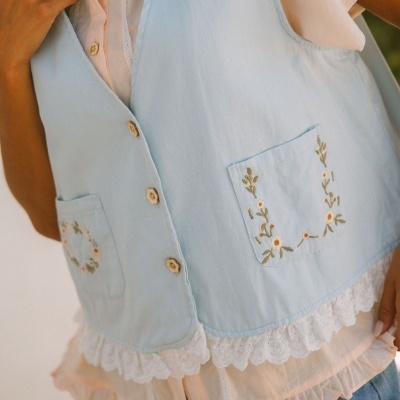 Colete azul claro bordado com flores e botões de madeira sobre camisa branca com renda