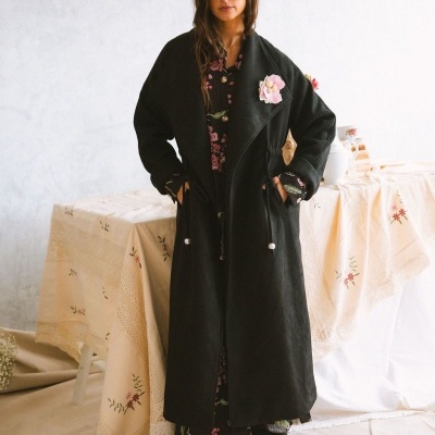 Mulher com casaco preto comprido e vestido floral fundo preto, sapatos castanhos.