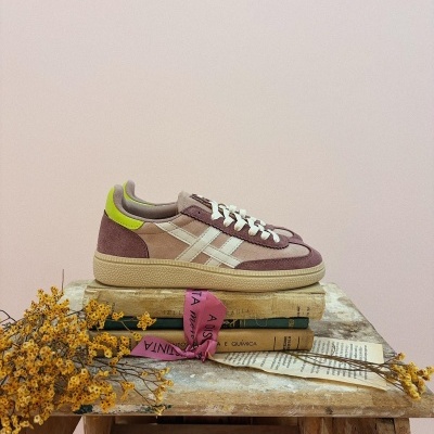 Ténis bege e castanho com riscas brancas e detalhe amarelo sobre livros antigos e banco de madeira com flores secas.