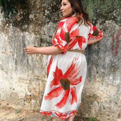 Mulher com vestido midi branco com grandes estampas florais vermelhas e castanhas, usando cinto bege e sandálias castanhas, em fundo de parede de pedra e plantas.