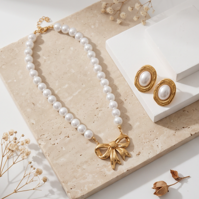 Colar de pérolas brancas com pendente dourado laço e brincos dourados com centro branco sobre base de pedra bege.