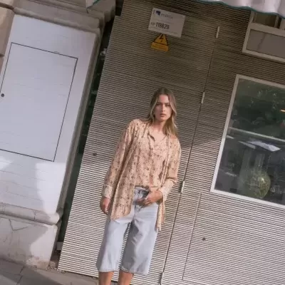 Mulher com blusa bege floral, calças denim curtas e sandálias pretas, em rua urbana com parede metálica e toldo.