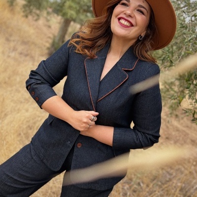 Mulher sorridente com blazer cinza escuro com costura laranja, calças cinza escuro e chapéu castanho em ambiente natural