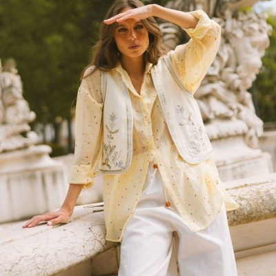 Mulher vestindo camisa amarela com colete branco bordado e calças brancas num jardim