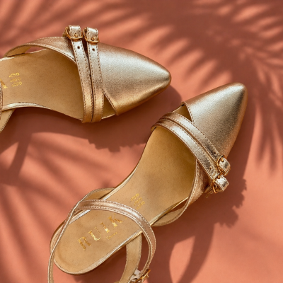 Sapatos dourados de salto com fivelas em fundo castanho e sombra de plantas