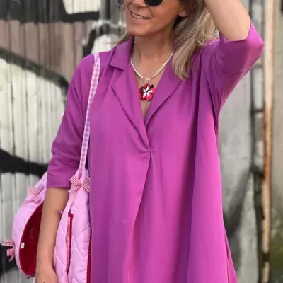 Pessoa com vestido rosa e mochila rosa, óculos de sol redondos pretos, colar de pérolas e pingente de flor
