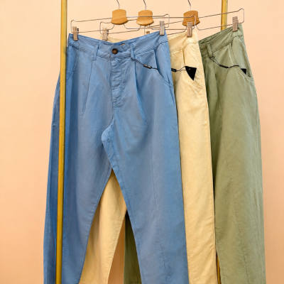 Três calças azul claro, creme e verde-oliva penduradas em cabides de madeira num varão amarelo