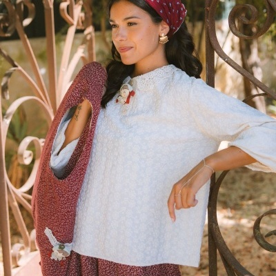 Mulher com blusa branca de padrão floral e saia vermelha de pequenas flores, com lenço de cabeça vermelho, em ambiente exterior com estrutura de ferro