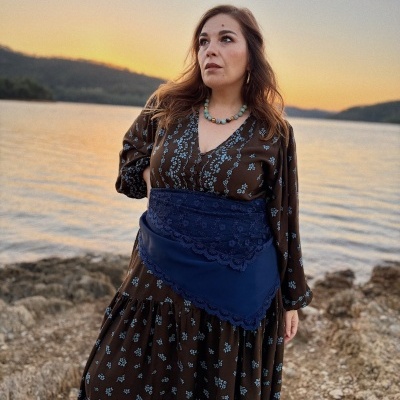 Mulher com vestido castanho com padrão floral azul e cinto azul largo junto a um lago ao pôr do sol