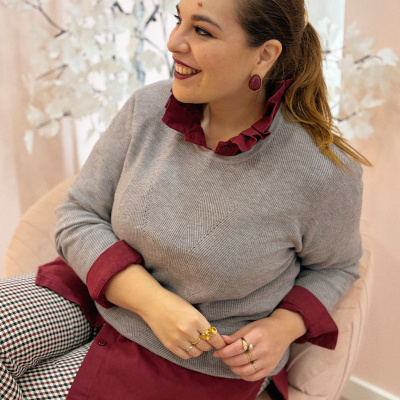 Mulher sorridente sentada usando blusa cinzenta, camisa bordô e calças xadrez, com brincos bordô e anéis dourados num ambiente com flores brancas.
