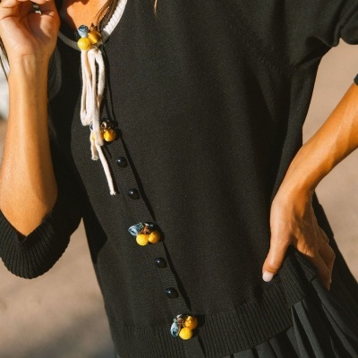 Camisola preta com botões e detalhes amarelos e azuis, gola branca com cordões, usada com saia preta plissada.