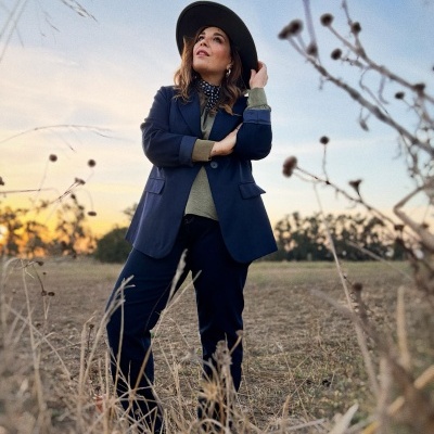 Mulher com casaco azul, calças azuis e chapéu preto em campo ao entardecer