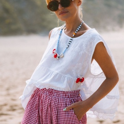Mulher com blusa branca bordada, calças vermelhas xadrez e óculos de sol na praia.