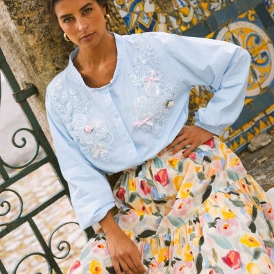 Blusa azul clara bordada com detalhes florais e laços, com saia floral colorida, em cenário exterior com azulejos