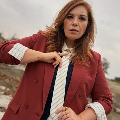 Mulher com blazer vermelho, camisa branca riscada, colete azul escuro e gravata branca riscada