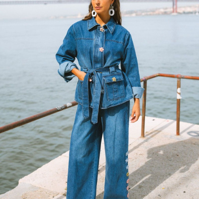 Mulher com conjunto azul de ganga com flores coloridas, à beira rio com ponte ao fundo