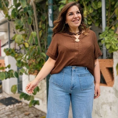 Mulher com blusa castanha e calças de ganga azuis posando com plantas ao fundo
