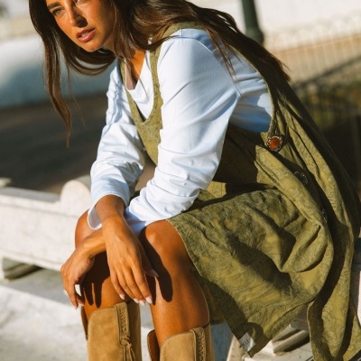 Mulher com vestido caqui, camisa branca e botas bege sentada ao sol
