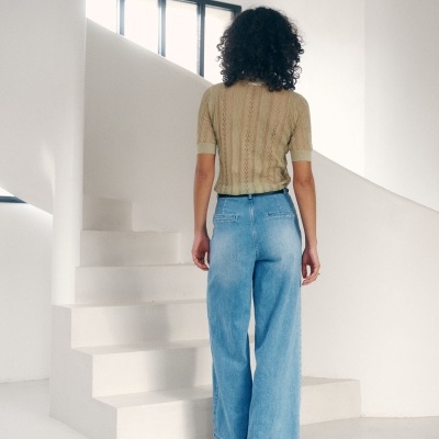 Pessoa de costas com camisola bege de malha e calças largas de ganga azul em ambiente branco com escadas.