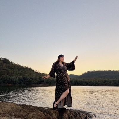 Mulher com vestido comprido preto com padrão floral junto a lago ao pôr do sol