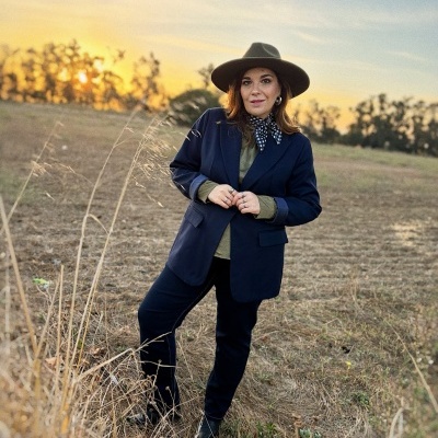 Mulher vestindo fato azul-escuro, lenço xadrez e chapéu castanho em campo ao pôr do sol
