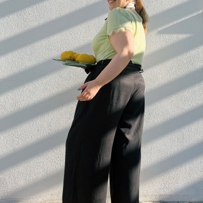 Mulher com camisa verde clara e calças pretas largas segurando prato com limões diante de parede branca.